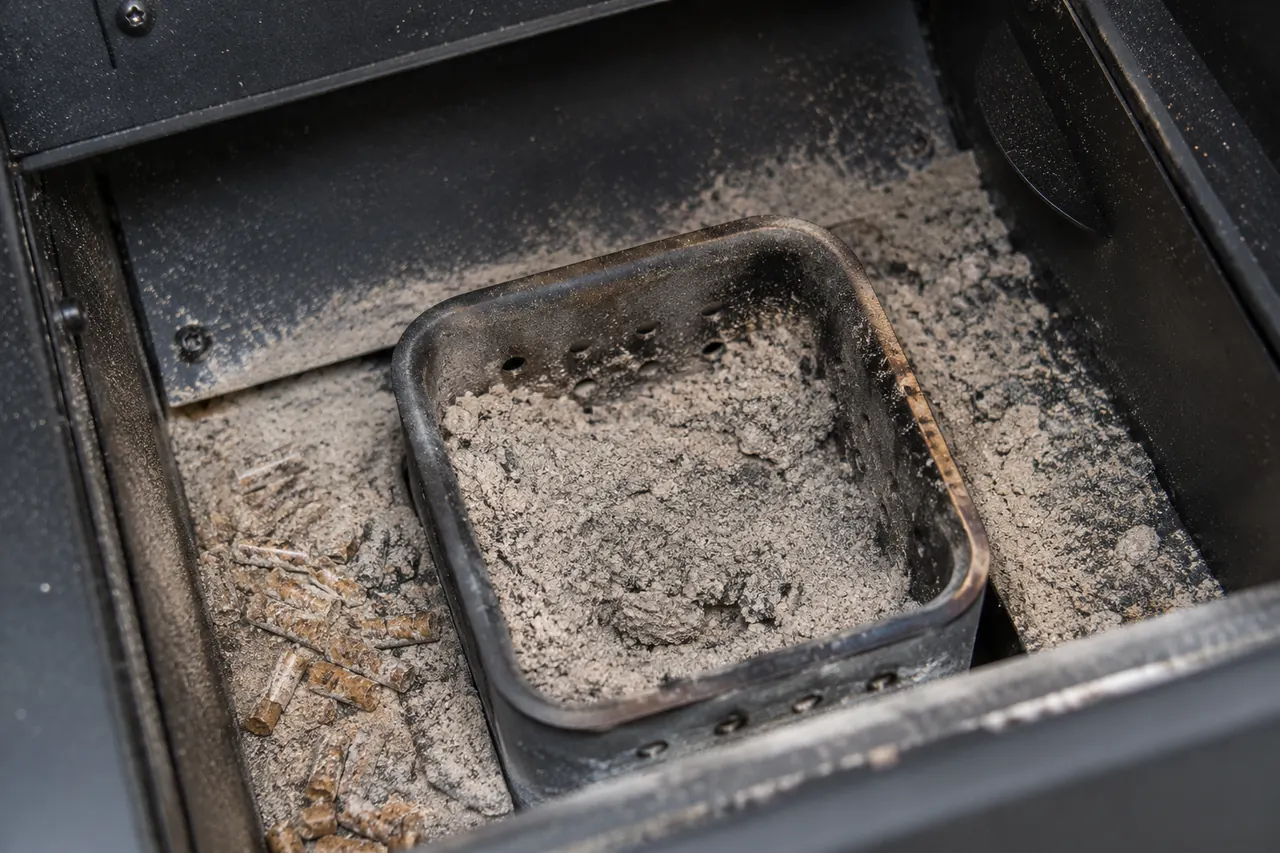 A cool pellet grill fire pot with a heavy buildup of ash and a few unburned pellets nearby.